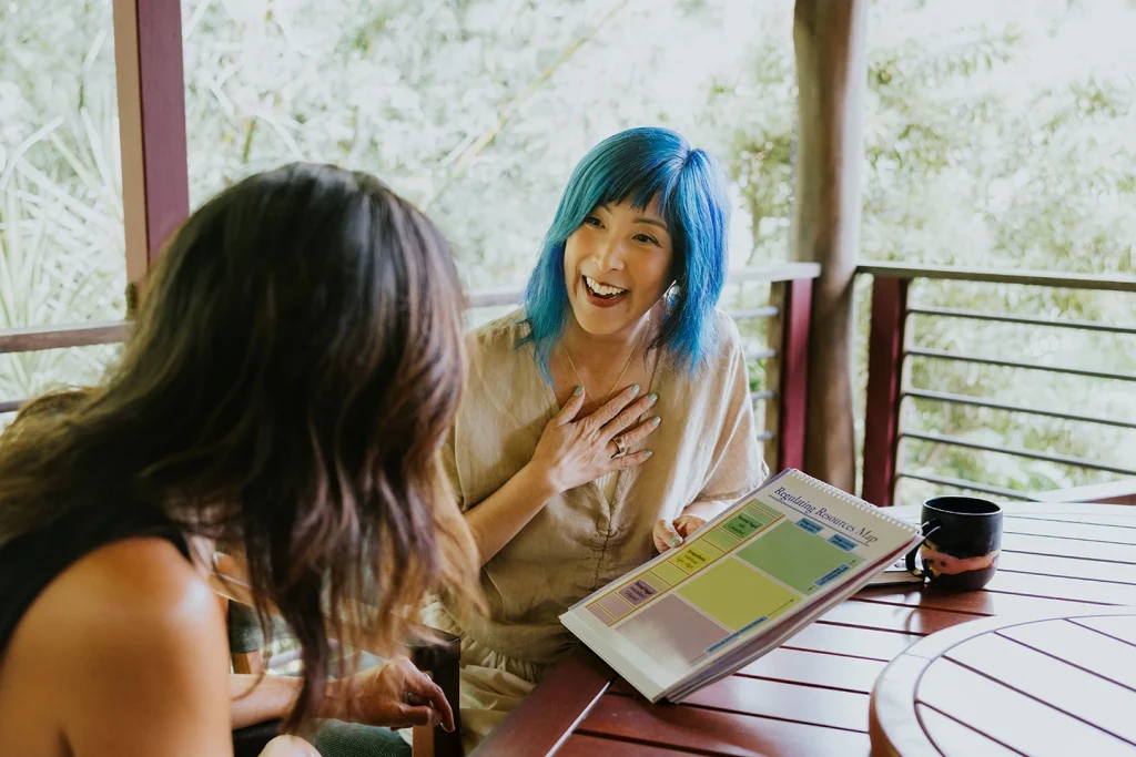 Jenn sitting at a table talking with a client. Jenn is holding her right hand to her heart and is holding a flipchart in her left hand.